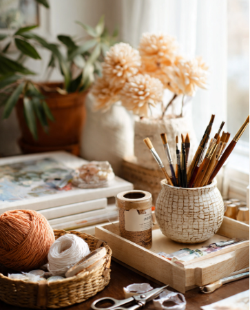 Flat lay of essential craft tools on rustic wood table including scissors, glue gun, washi tape, yarn spools, paint brushes, and colored paper in neutral boho aesthetic