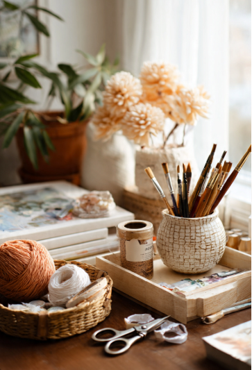 Flat lay of essential craft tools on rustic wood table including scissors, glue gun, washi tape, yarn spools, paint brushes, and colored paper in neutral boho aesthetic