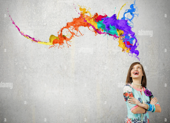 A smiling woman with arms crossed looking up at a vibrant, colorful splash of paint bursting across a gray wall, illustrating why is beauty important in life."