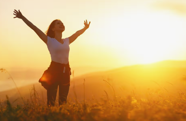 "A joyful woman standing on a hilltop with her arms raised wide, welcoming the warm glow of a brilliant sunset to illustrate why is beauty important in life."