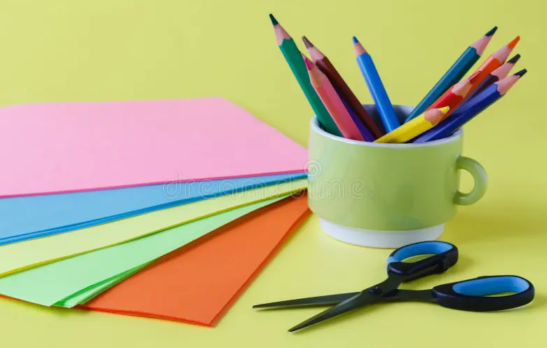 A stack of colorful construction paper, a pair of scissors, and a green mug filled with colored pencils.