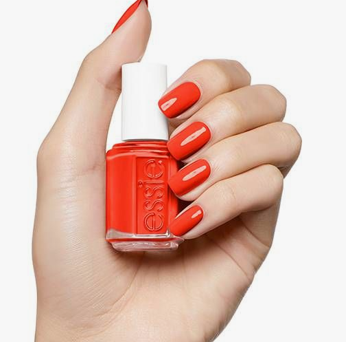 What nail colour is trending now demonstrated as vibrant, glossy orange-red polish on short square nails holding an Essie nail polish bottle.