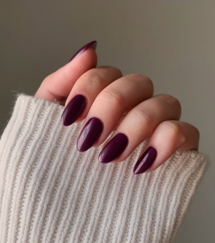 What nail colour is trending now showcased as a glossy, deep dark plum or burgundy shade on oval-shaped nails against a white knit sleeve.