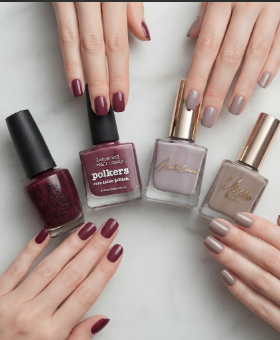 A flatlay featuring four nail polish bottles in rich berry, plum, and taupe shades alongside beautifully manicured hands showcasing matching glossy nail colors on a clean white marble background.