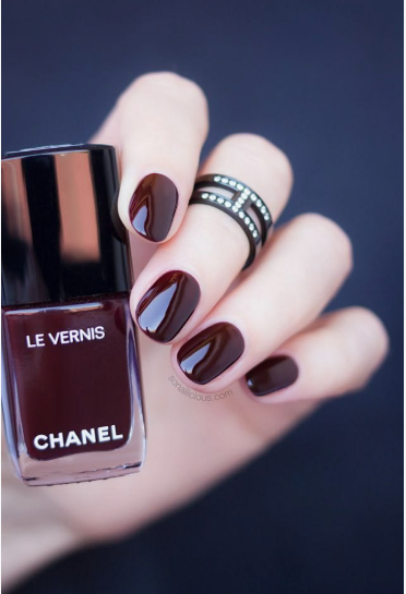 A hand holding a bottle of Chanel Le Vernis dark burgundy nail polish to demonstrate nail colors that age your hands.