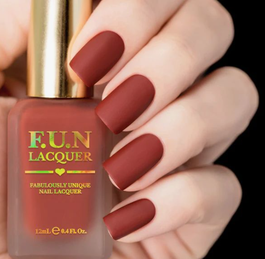 A hand holding a bottle of F.U.N Lacquer matte rust-red nail polish in a discussion about nail colors that age your hands.