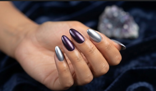 A hand with almond-shaped nails featuring a combination of glossy metallic purple and cool grey nail polish, illustrating nail colors that age your hands.