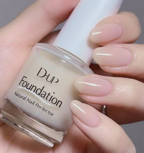 A hand holds a bottle of D-UP Foundation Natural Nail Perfector, showcasing manicured fingernails painted with a glossy, sheer neutral polish.