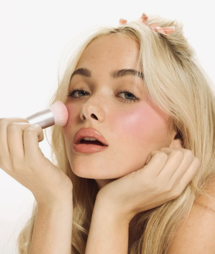 10 Makeup Trends for 2026: The Beauty Looks Everyone Will Want to Try A fair-haired model demonstrating makeup trends 2026 by applying a soft pink blush to her cheekbone with a small brush, featuring glossy lips and a natural, dewy complexion.