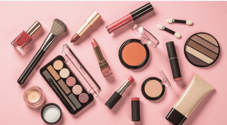 A flatlay arrangement of various cosmetics and beauty tools on a pink background, featuring eyeshadow palettes, lipsticks, compact powders, makeup brushes, nail polish, and foundation tube in line with makeup trends 2026.