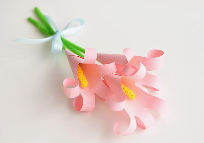 15 Easy DIY Paper Flowers for Kids: Fun Crafts for Toddlers, Preschoolers & Older Children A small bouquet of handmade light pink paper lily flowers with curled petals and yellow pipe cleaner stamens on green stems, tied together with a light blue ribbon bow, presented as easy paper flowers for kids.
