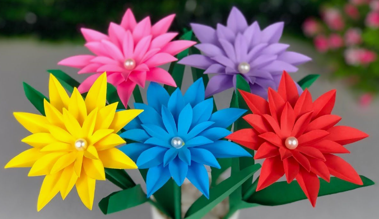A bouquet of vibrant multi-layered paper flowers in yellow, pink, blue, purple, and red, each featuring a pearl bead center and mounted on green stems with leaves, presented as easy paper flowers for kids.