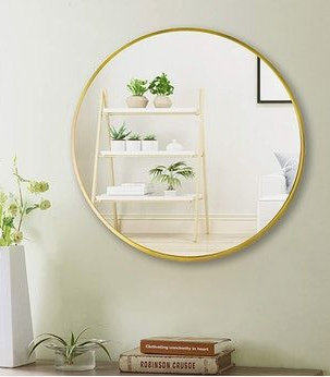 10 Cheap Ways to Make Your Home Look More Expensive A round wall mirror with a thin gold metal frame reflecting a shelf with potted plants and books, displayed as an example of cheap ways to make your home look more expensive.