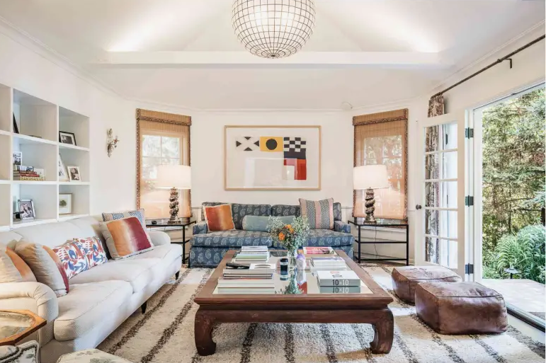 10 Cheap Ways to Make Your Home Look More Expensive A bright, sunlit living room featuring two patterned sofas, a mirrored wooden coffee table, built-in shelving, leather poufs, and open French doors leading to a lush green outdoor view, showcased as an example of cheap ways to make your home look more expensive.