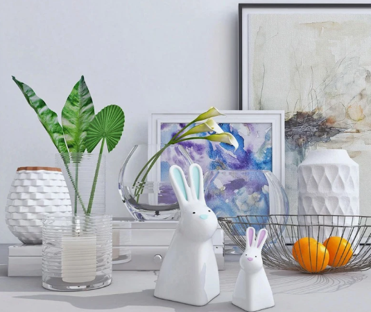 10 Cheap Ways to Make Your Home Look More Expensive A tabletop arrangement featuring ceramic white vases, potted green leaves, glass bowls with flowers, two white ceramic bunny figurines with blue and pink accents, a wire basket with oranges, and framed artwork in the background, presented as an example of cheap ways to make your home look more expensive.