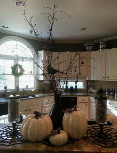 A striking arrangement featuring a dark vase holding tall bare branches with perched crows, surrounded by white pumpkins and glass apothecary jars on spiderweb placemats as stylish Halloween decor ideas for indoor spaces.