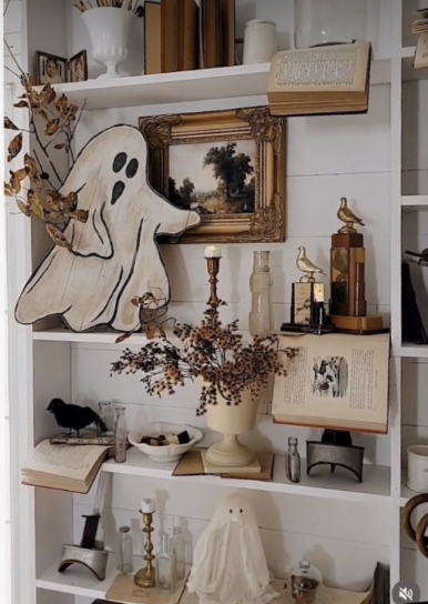 A white wooden ghost cutout resting beside a framed landscape painting on a white bookshelf, showcasing creative Halloween decor ideas for indoor spaces.