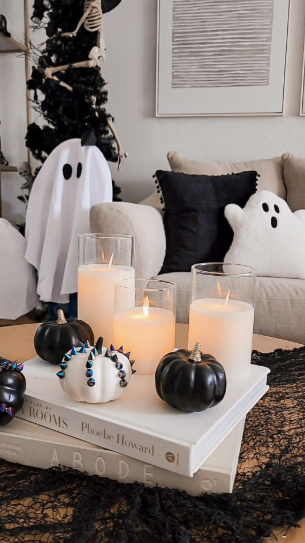 Stacked decorative coffee table books topped with pillar candles in glass cylinders and small black and white pumpkins, creating chic Halloween decor ideas for indoor spaces.