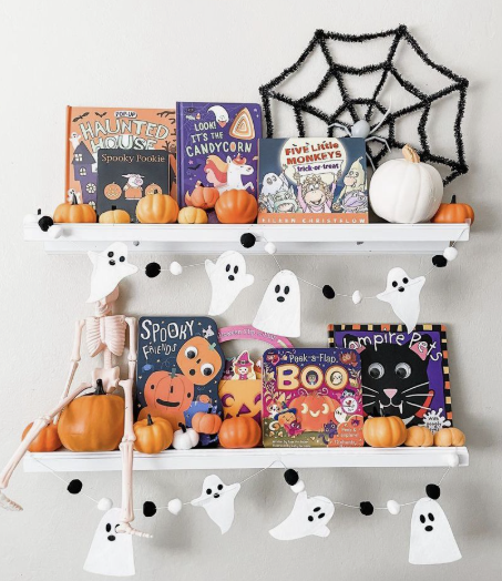 Two white floating shelves styled with colorful children's holiday books, miniature pumpkins, and a black tinsel spiderweb, creating playful Halloween decor ideas for indoor spaces.