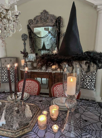 A tall black pointed witch hat trimmed with dark feathers perched atop a glass candle hurricane, serving as a striking focal point for Halloween decor ideas for indoor spaces.