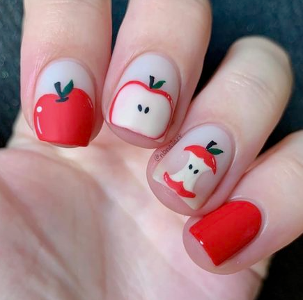 18 Back-to-School Nail Designs for a Fresh Start This School Year Close-up of charming back-to-school nail designs featuring cute red apple motifs, including whole apples, sliced apple halves with seeds, and an eaten apple core against a neutral base.