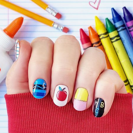 Close-up of colorful back-to-school nail designs featuring short, rounded nails painted with playful school-themed art, including blue stripes, a red apple, yellow and pink color-blocking, and an ABC chalkboard design, set against a background of pencils, crayons, and notebook paper.