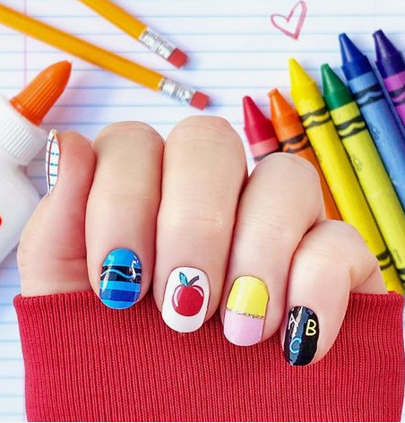18 Back-to-School Nail Designs for a Fresh Start This School Year Close-up of colorful back-to-school nail designs featuring short, rounded nails painted with playful school-themed art, including blue stripes, a red apple, yellow and pink color-blocking, and an ABC chalkboard design, set against a background of pencils, crayons, and notebook paper.