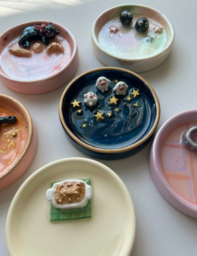 A group of colorful small dishes featuring fun air dry clay ideas, including trinket trays with miniature cats, celestial star patterns, and cute sculpted food.