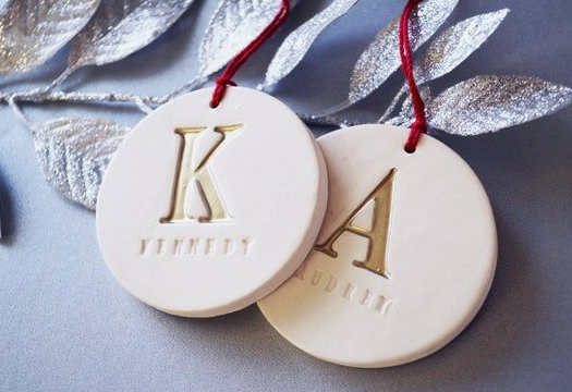 Two custom round white ornaments showcasing personalized air dry clay ideas, featuring gold-accented embossed initials and names hanging on red string against a silver background.