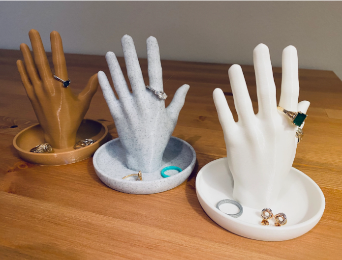 Three sculpted hand-shaped jewelry holders demonstrating creative air dry clay ideas, featuring brown, speckled grey, and white hands rising from round trinket dishes to hold rings and accessories.