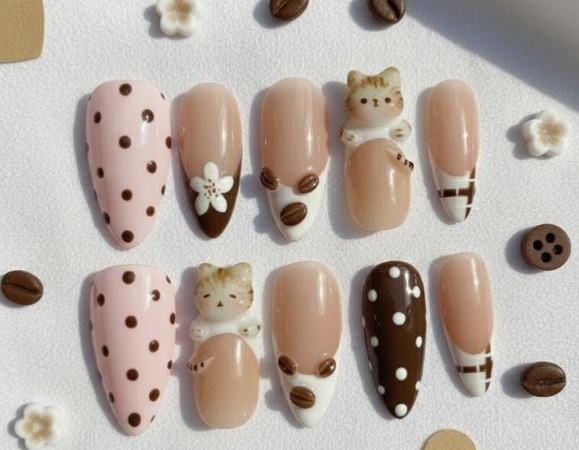 A set of ten press-on nails featuring a cozy coffee and cat theme in shades of brown, cream, and pink, displayed alongside coffee bean charms and small white flowers.