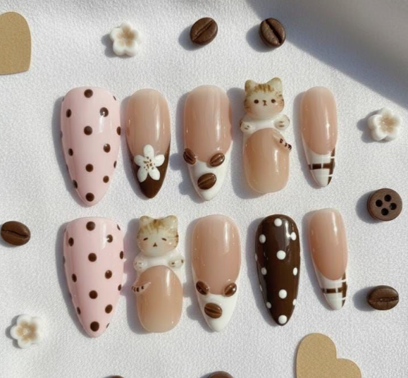 15 Stunning 3D Nail Designs to Try This Season A set of ten press-on nails featuring a cozy coffee and cat theme in shades of brown, cream, and pink, displayed alongside coffee bean charms and small white flowers.
