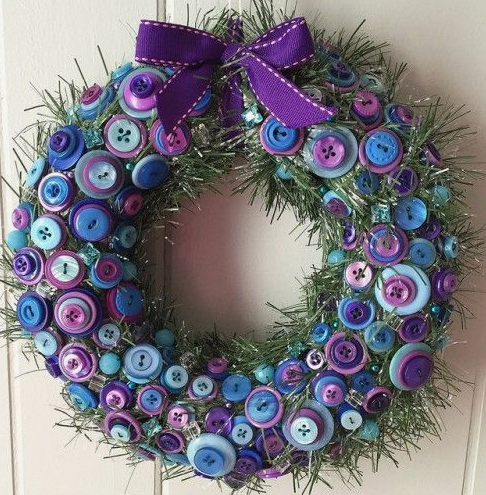 30 Creative DIY Button Crafts to Turn Spare Buttons Into Beautiful Decor A holiday wreath decorated with tinsel and an abundance of blue and purple buttons, accented with a purple ribbon bow, highlighting DIY button crafts.