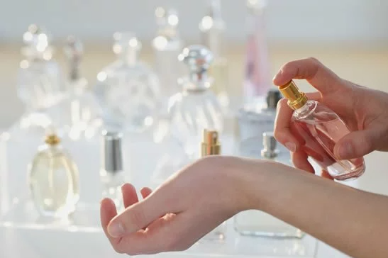 Close-up of a person spraying perfume onto their wrist surrounded by a collection of fragrance bottles, representing Fall 2026 Beauty Trends.
