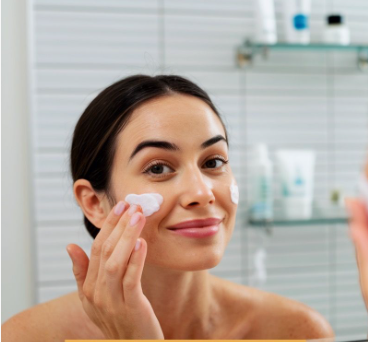 A close-up shot of a smiling woman gently applying a white moisturizing cream to her cheeks in front of a bathroom mirror as part of a low maintenance beauty routine.