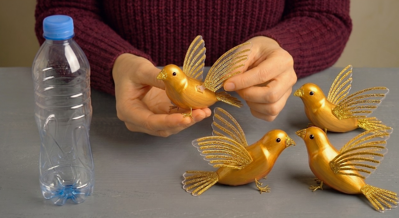 A DIY craft project showing gold-painted decorative birds with glittery wings made from recycled plastic bottles, displayed alongside an empty plastic bottle.