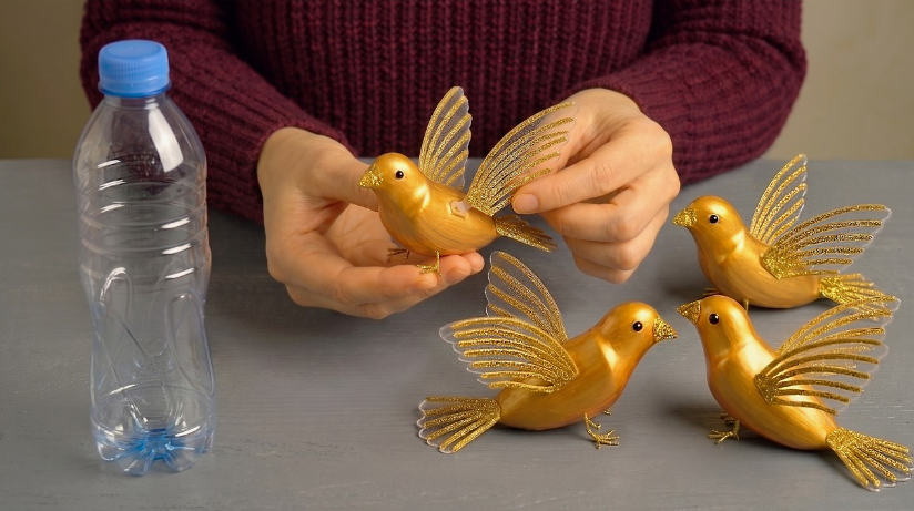 A DIY craft project showing gold-painted decorative birds with glittery wings made from recycled plastic bottles, displayed alongside an empty plastic bottle.