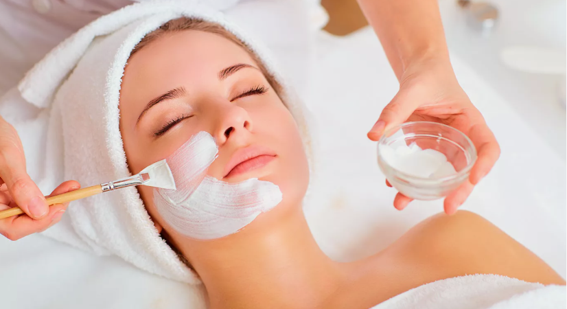 A spa professional using a brush to apply a white cream mask to a relaxed woman's face from a small glass bowl, demonstrating a professional treatment that contrasts with a simple skincare routine for beginners.