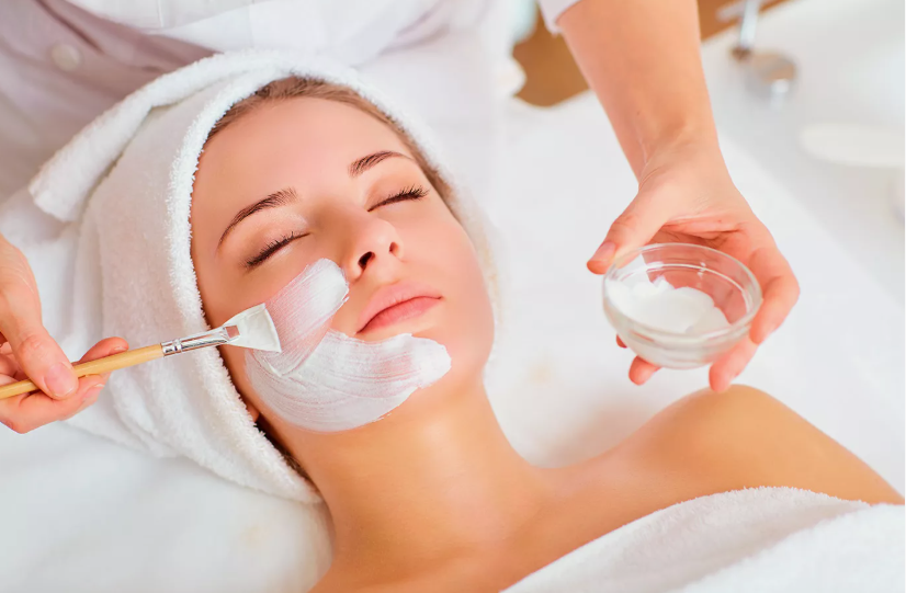 A spa professional using a brush to apply a white cream mask to a relaxed woman's face from a small glass bowl, demonstrating a professional treatment that contrasts with a simple skincare routine for beginners.