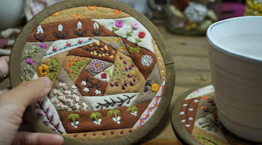 A hand-stitched, round fabric mug rug featuring a patchwork design in earthy tones decorated with intricate floral and botanical embroidery, presented as a cozy handmade project under the keyphrase one hour crafts.
