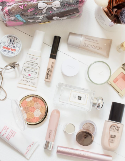 Beauty Products You Actually Need: A Minimalist Guide to Your Beauty Collection Flat lay arrangement of essential beauty products you actually need, including foundation, liquid blush, concealer, lipstick, and perfume bottles on a white wooden surface.