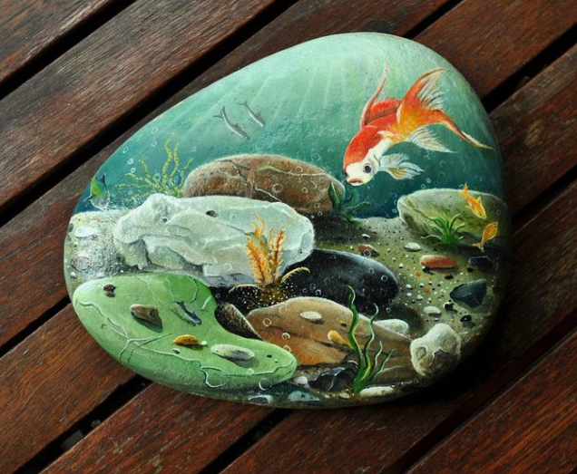A large, smooth garden stone meticulously hand-painted with an underwater aquatic scene featuring a vibrant goldfish, river rocks, and aquatic plants, showcasing stunning home decor ideas without buying anything.