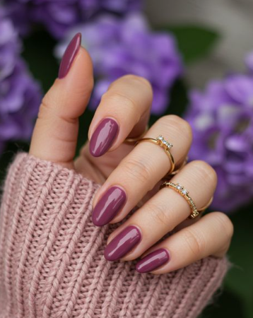 Hands showcasing classy nail colors for mature hands, featuring glossy mauve-purple oval nails accessorized with delicate diamond rings and a cozy knit sweater against purple hydrangeas.