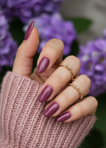 Hands showcasing classy nail colors for mature hands, featuring glossy mauve-purple oval nails accessorized with delicate diamond rings and a cozy knit sweater against purple hydrangeas.