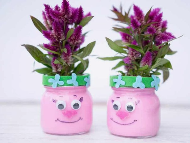 "Two pink painted glass jar vases decorated with googly eyes, smiling faces, and green felt bands, holding vibrant purple flowers as part of 25 easy DIY home decor crafts for kids' rooms."