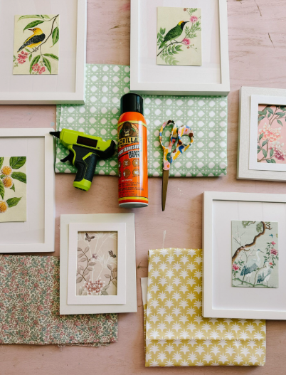 A flat lay showing white picture frames, fabric pieces, scissors, a glue gun, and adhesive spray arranged for upcycling projects, showcasing creative DIY crafts with old clothes.