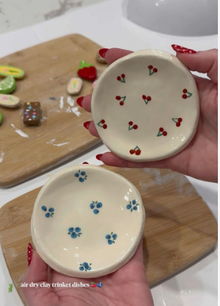 A close-up view of two handmade air dry clay crafts shaped into small round trinket dishes, one painted with red cherries and the other with blue blueberries.