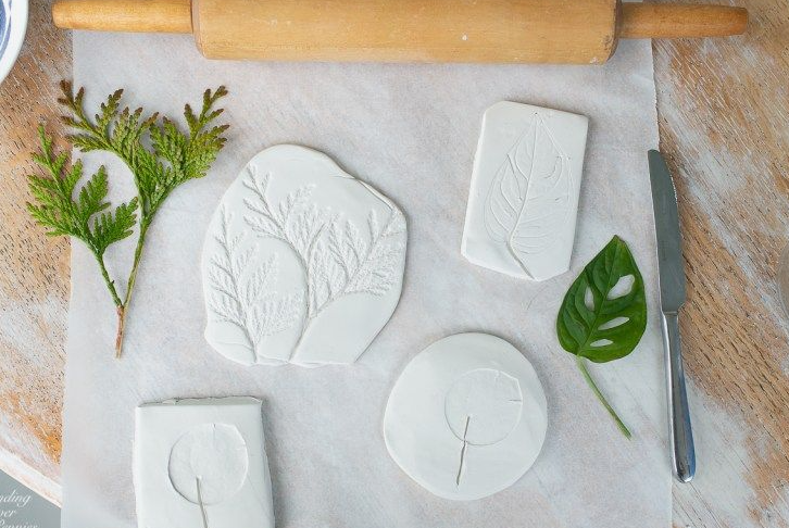 An overhead view of process shots for air dry clay crafts, featuring white clay pieces pressed with real leaves and ferns on parchment paper alongside a rolling pin, knife, and plant sprigs.