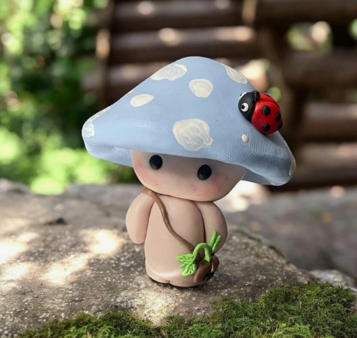 A close-up of a whimsical air dry clay craft shaped into a cute mushroom person with a blue-and-white spotted cap, a ladybug resting on top, and holding a tiny green sprout.