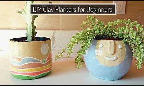 Two handmade air dry clay crafts shaped into colorful planters holding succulents, featuring a cylindrical pot painted with a scenic landscape and a round pot sculpted with a smiling face.